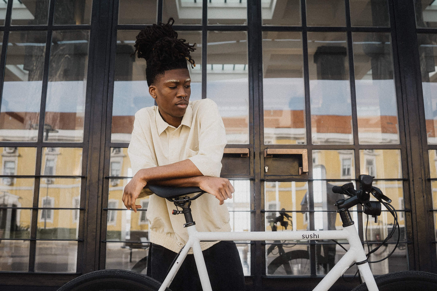 Eine Person lehnt sich entspannt auf den Sattel eines weißen SUSHI Bikes. Im Hintergrund ist ein Gebäude mit großen Fenstern und gelber Fassade zu sehen.