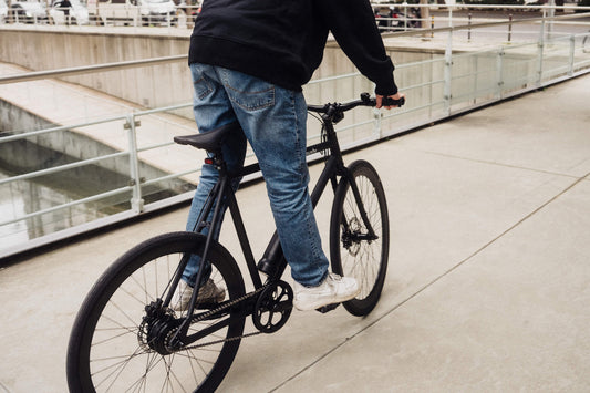 Person fährt ein schwarzes E-Bike von SUSHI BIKES auf einem modernen Gehweg mit Glasgeländer.
