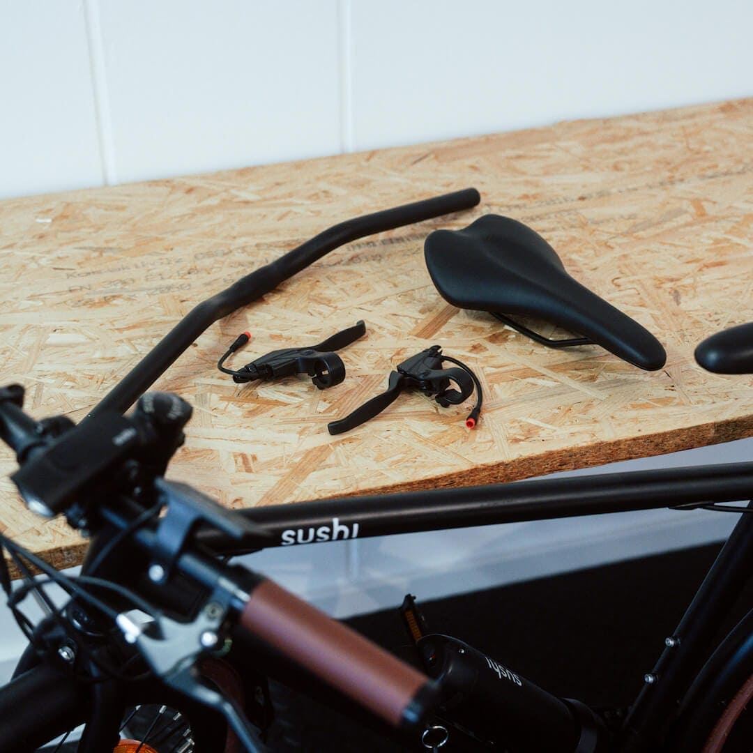 Ergo-Lenker+, Sattel des Maki+ und Bremshebel+ auf einer Holzplatte liegend neben einem SUSHI BIKE