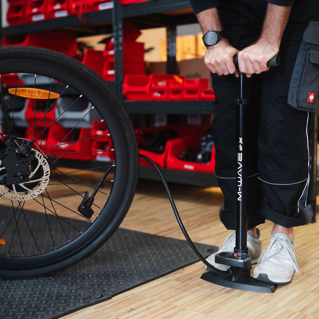 Person pumpt den Hinterreifen eines SUSHI BIKES mit der Standpumpe in einer Werkstatt auf, beide Füße stehen fest auf der rutschfesten Trittplatte