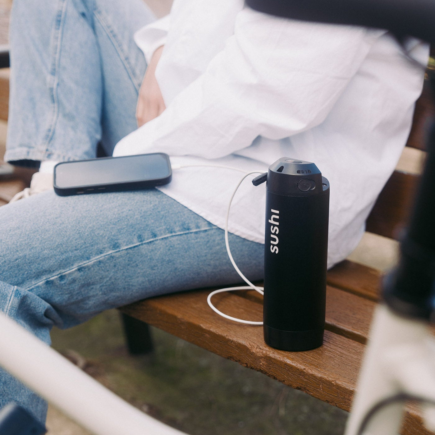 Akku eines SUSHI BIKE lädt ein Smartphone über USB, Person in Jeans sitzt entspannt auf einer Parkbank