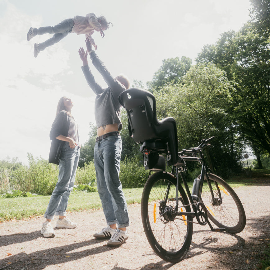 Familie mit Kind beim Spielen neben SUSHI BIKE mit montiertem Kindersitz, Hinweis auf Zubehör-Kompatibilität