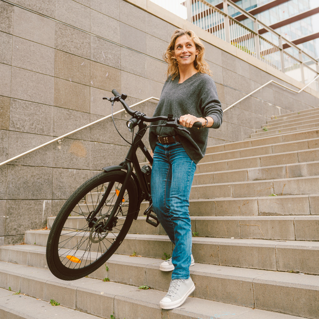 Frau trägt ein schwarzes E-Bike California Roll 3.0+ lächelnd eine Steintreppe herunter.