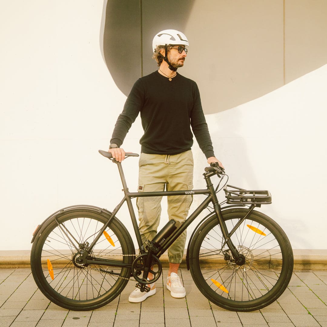 Maki 3.0+ Black wird am Lenker und Sattel von Mann mit weißem Helm vor weißer Hauswand gehalten