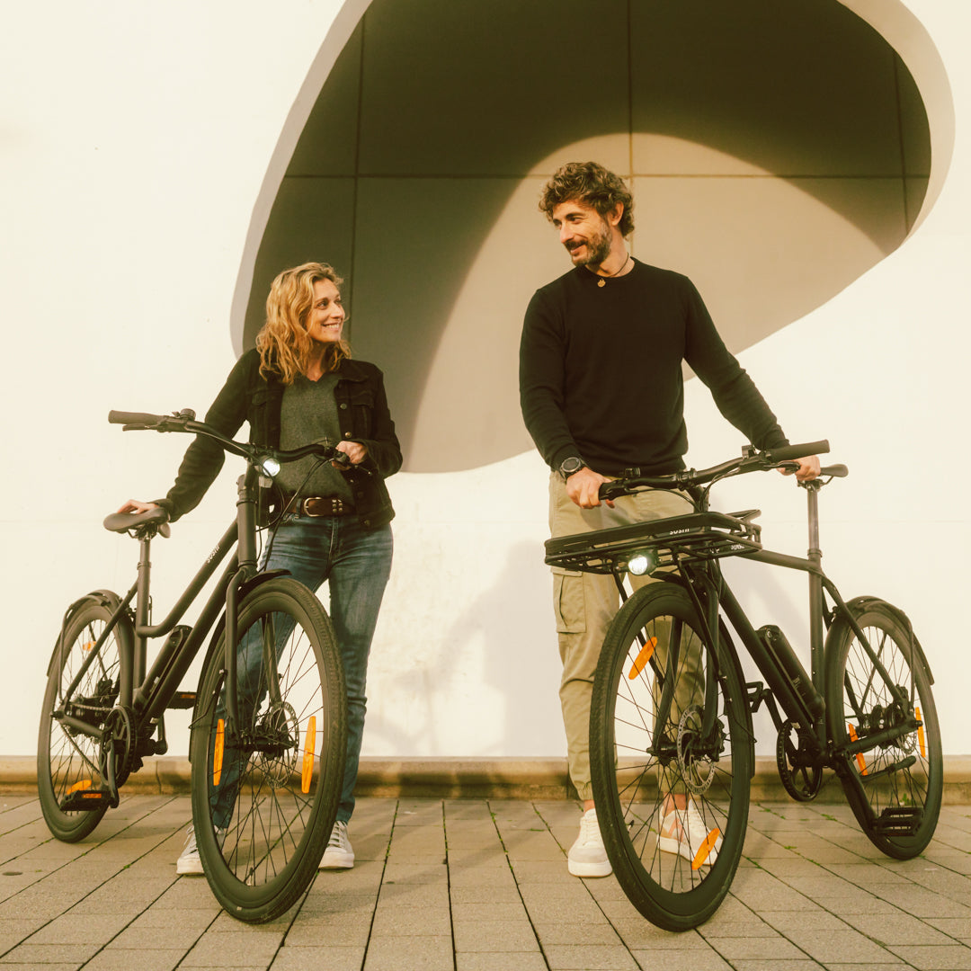 Zwei Personen halten schwarze SUSHI Bikes vor moderner Wand, beide lächeln sich an, während sie nebeneinander stehen