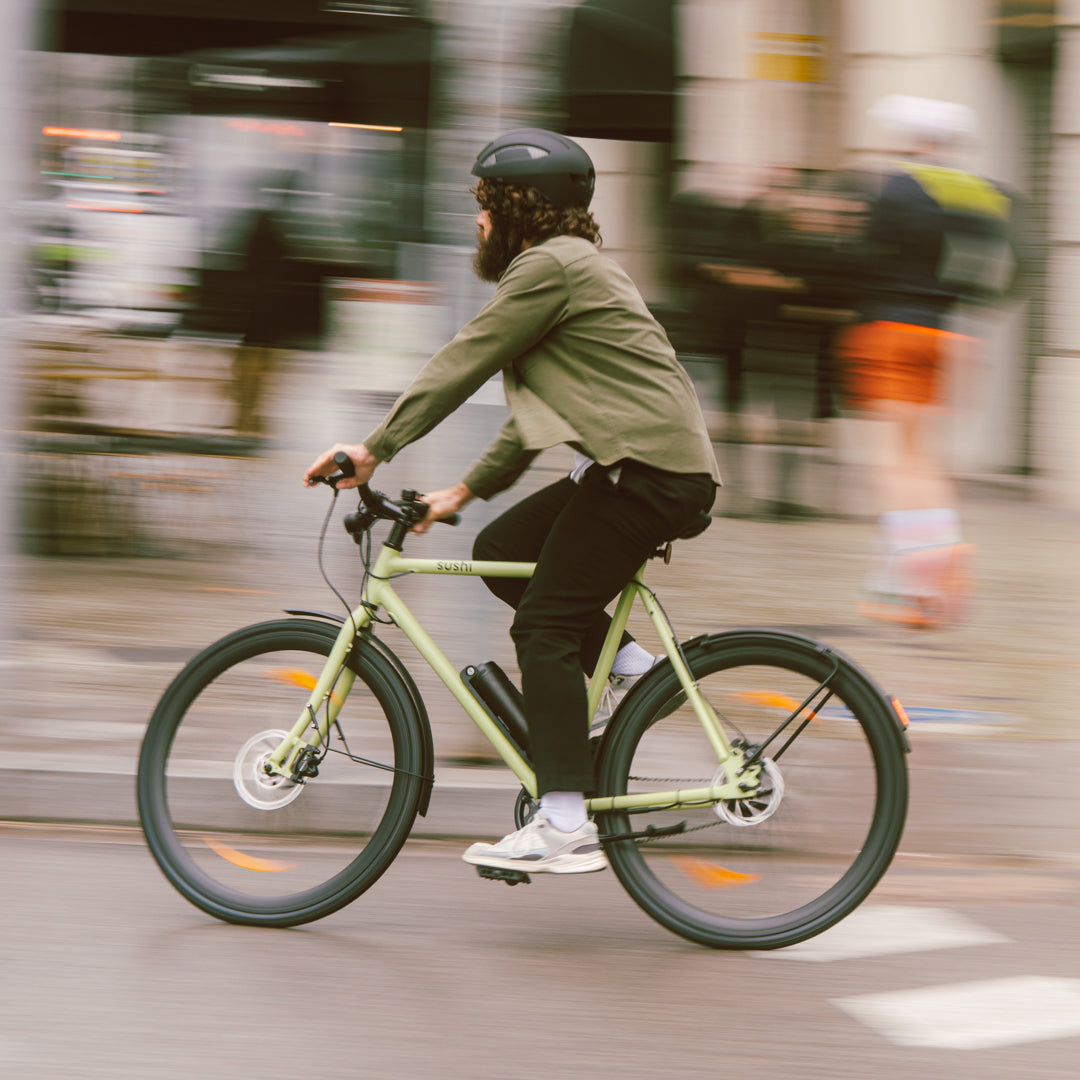 Person mit Helm fährt dynamisch das Maki 3.0+ E-Bike in der Farbe Matcha durch die Stadt
