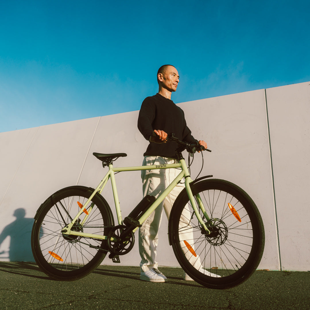 Mann mit SUSHI BIKES E-Bike Maki 3.0+ in Matcha vor weißer Wand bei Sonne