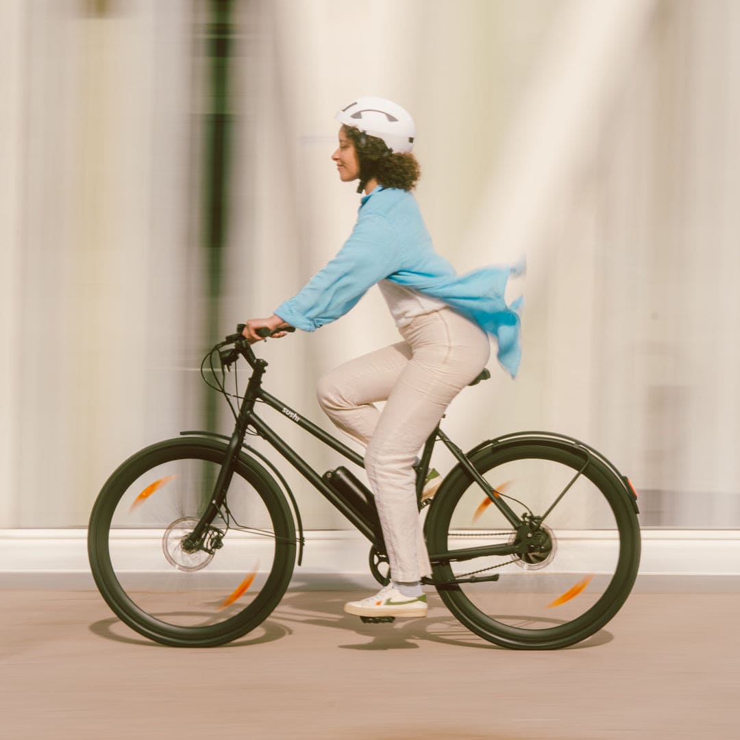 California Roll 3.0+ Black: Frau fährt schwarzes E-Bike mit weißem Helm und wehender blauer Bluse.