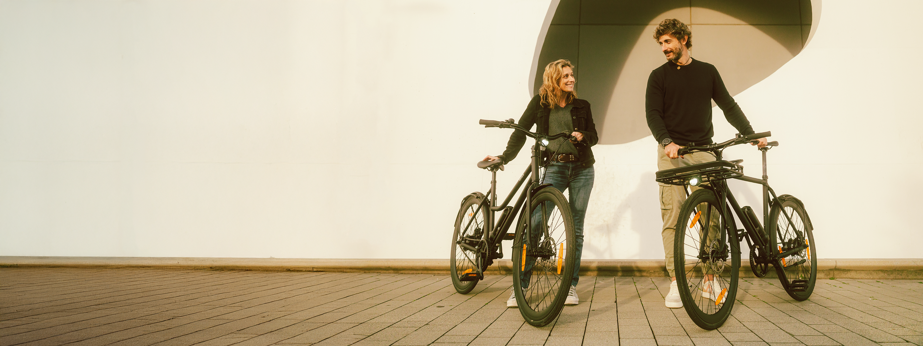 Frau und Mann stehen mit City E-Bike California Roll 3.0+ und Maki 3.0+ in schwarz vor Hauswand