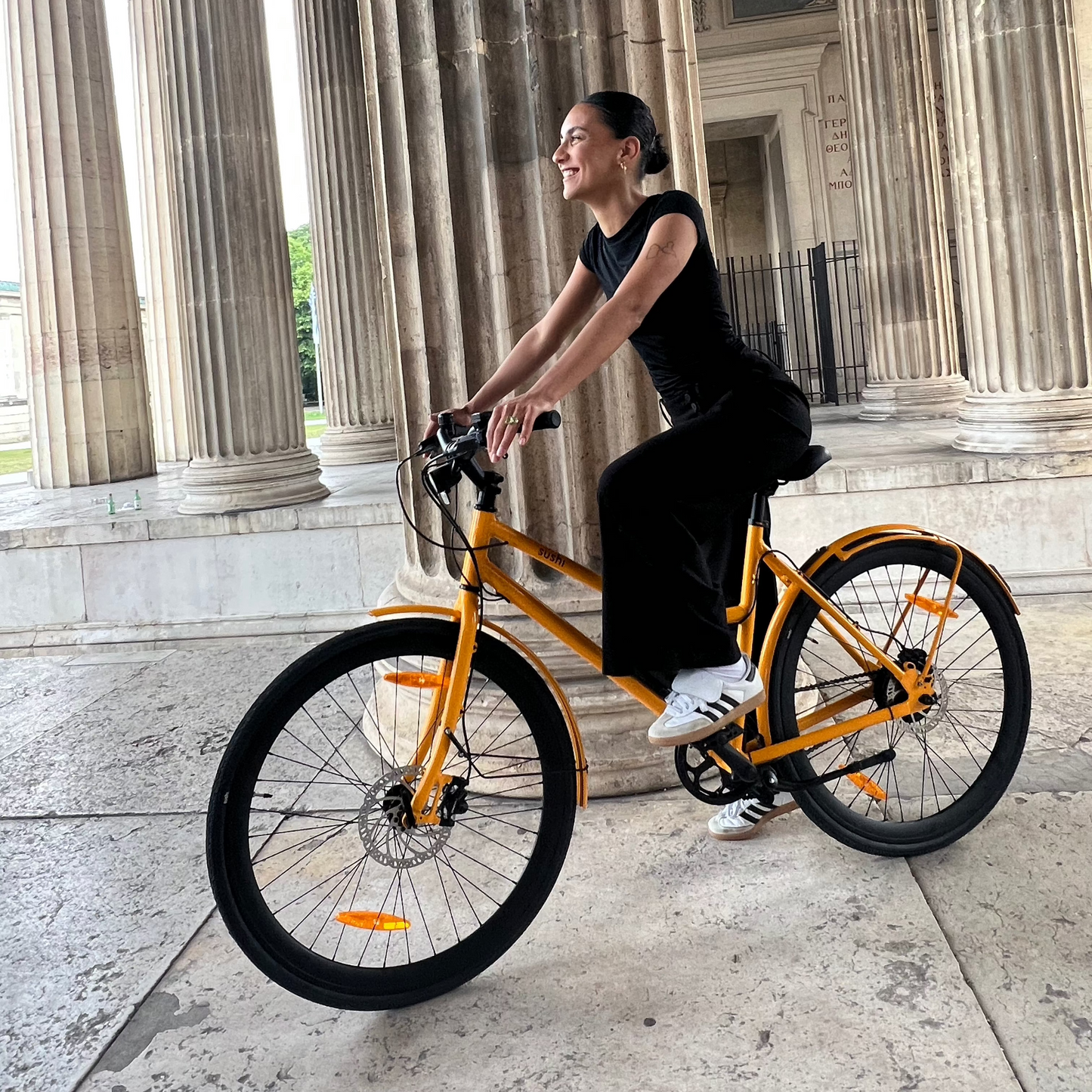 Frau sitzt auf E-Bike California Roll 3.0+ in Lava vor Säulenhalle im Sonnenschein