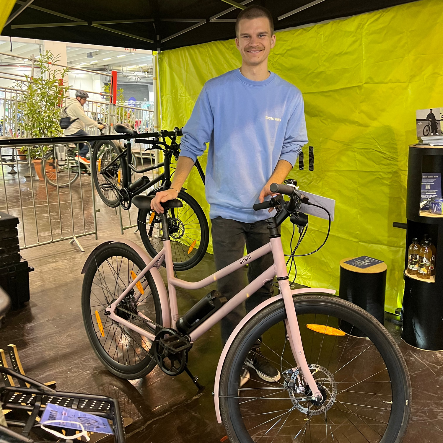 Produktmanager Laurin hält ein rosa SUSHI Bike an einem Messestand und lacht in die Kamera