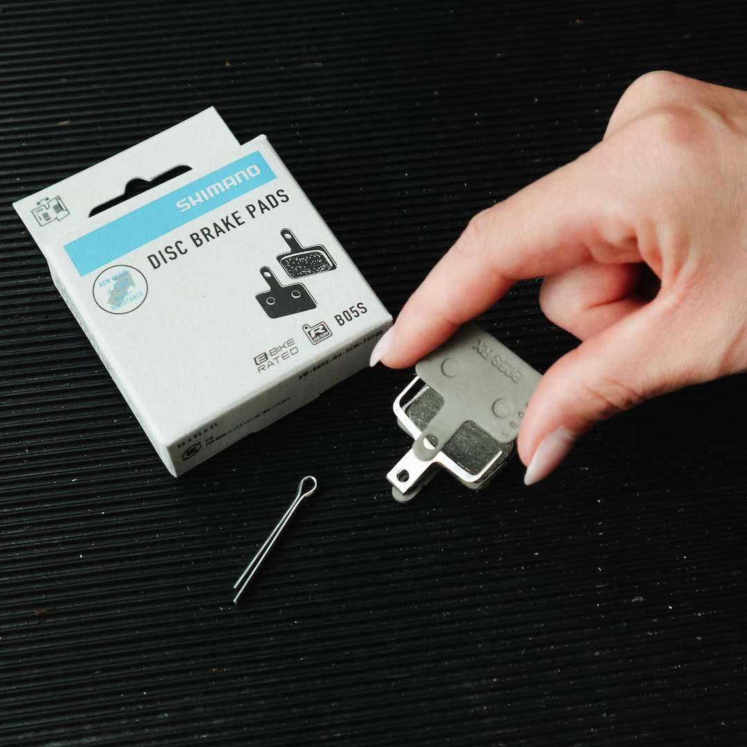 Hand hält Shimano Bremsbelag neben geöffneter Produktverpackung und Befestigungssplint auf schwarzer Unterlage