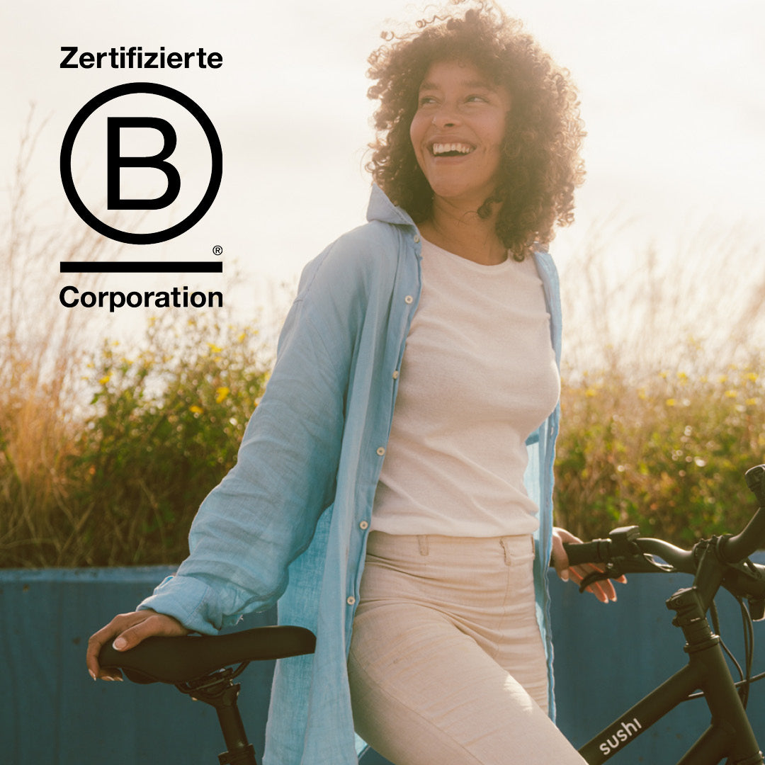 Lächelnde Frau steht mit schwarzem SUSHI E-Bike neben B Corp™-Logo im Sonnenlicht