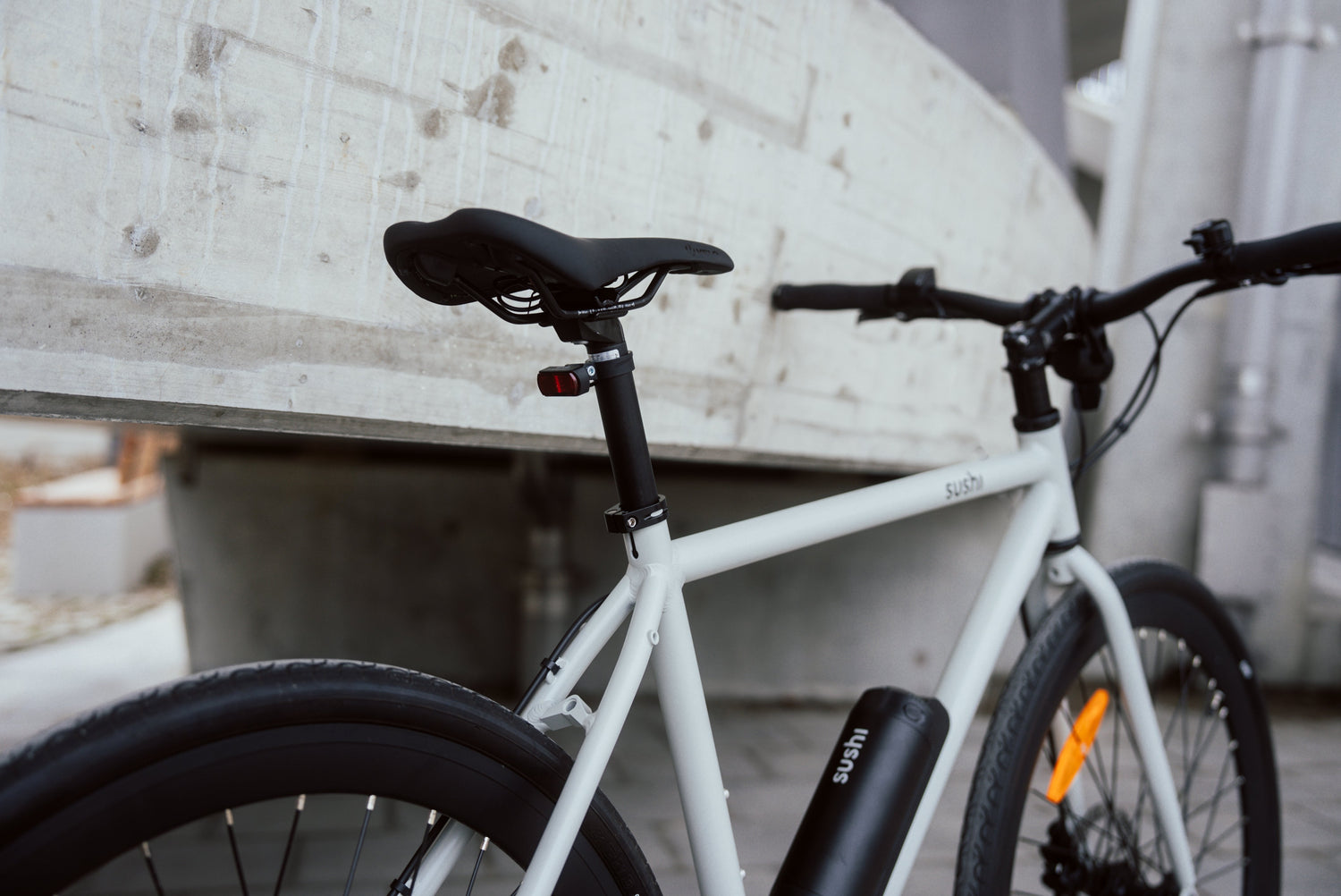 Graues SUSHI BIKES E-Bike angelehnt an der Wand. Im Fokus steht der Rahmen mit Akku und Sattel, im Hintergrund ist eine moderne Betonarchitektur erkennbar.