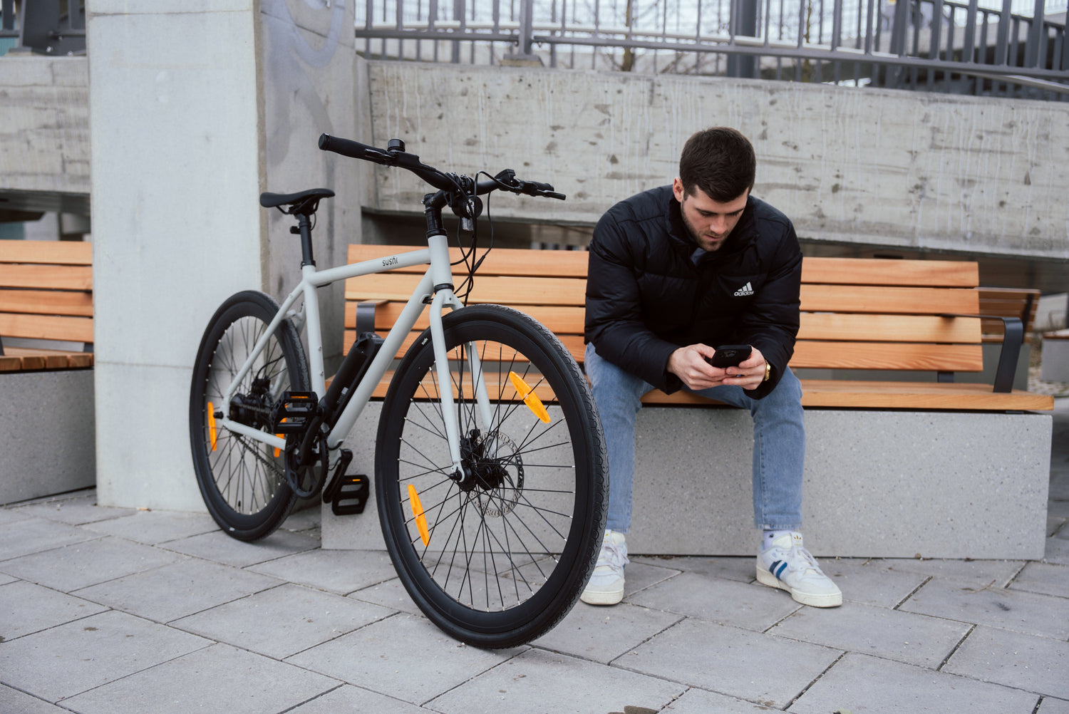 Ein junger Mann sitzt auf einer Bank und schaut auf sein Handy, neben ihm steht ein graues SUSHI BIKES 3.0 E-Bike.