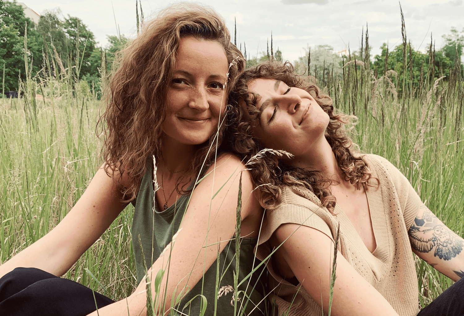 Zwei Frauen sitzen entspannt in hohem Gras und lächeln. Die natürliche Umgebung mit Bäumen und Pflanzen im Hintergrund vermittelt eine ruhige, sommerliche Atmosphäre.