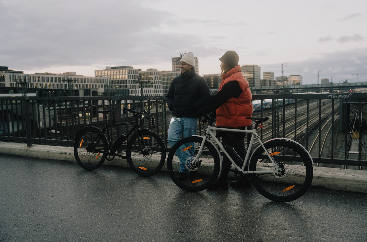 Zwei Personen mit SUSHI BIKES E-Bikes auf einer Brücke bei Stadtpanorama