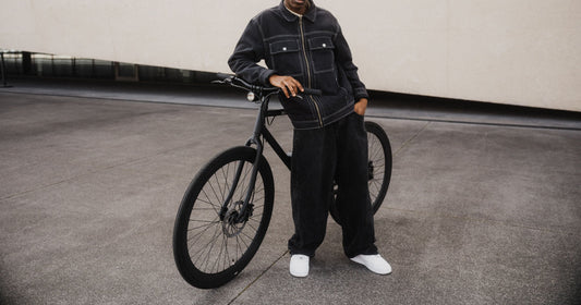 Person in schwarzem Outfit lehnt lässig an einem schwarzen SUSHI BIKES E-Bike vor moderner urbaner Architektur.