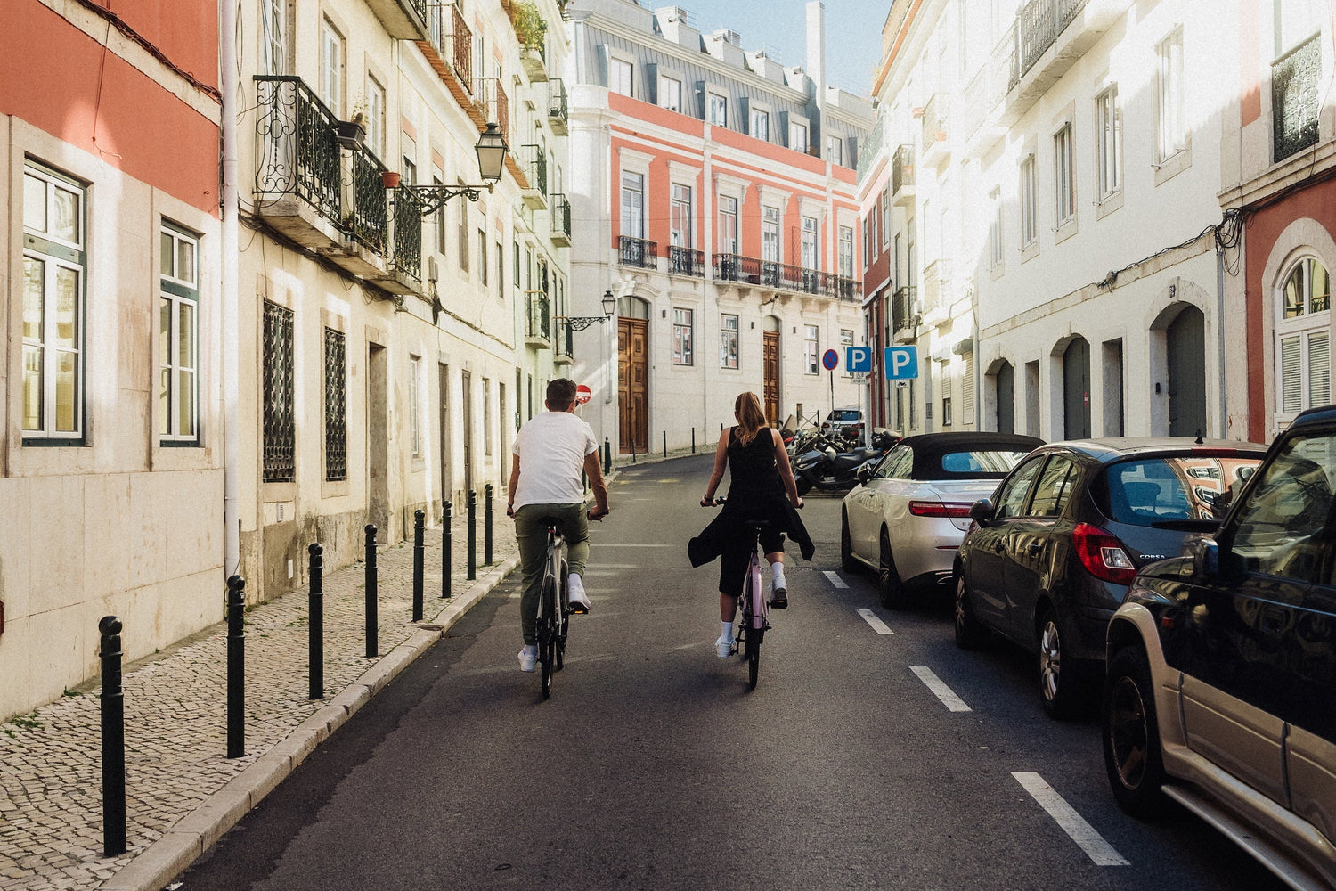 Auf dem Bild fahren zwei Personen mit dem Fahrrad durch eine sonnige, charmante Altstadtstraße mit parkenden Autos und historischen Gebäuden.
