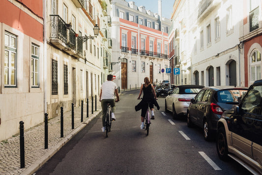 Auf dem Bild fahren zwei Personen mit dem Fahrrad durch eine sonnige, charmante Altstadtstraße mit parkenden Autos und historischen Gebäuden.