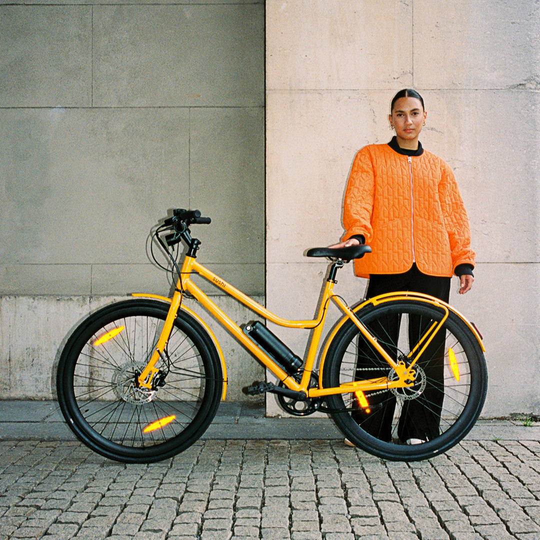 California Roll 3.0+ Lava, Frau in orangener Jacke steht hinter E-Bike mit tiefem Einstieg vor Betonwand 