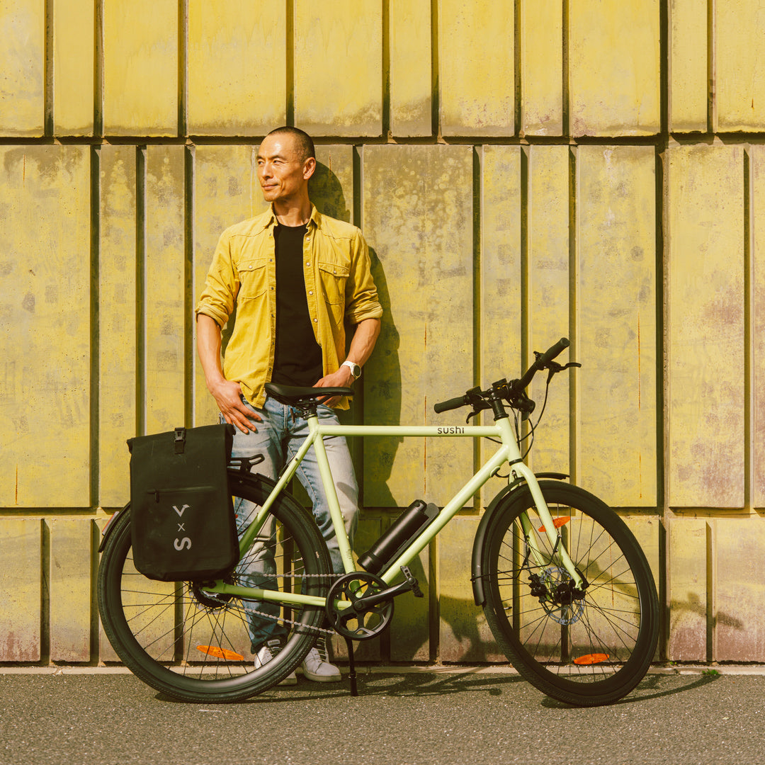 Maki 3.0+ Matcha, Mann steht mit E-Bike und Fahrradtasche vor einer gelben Wand