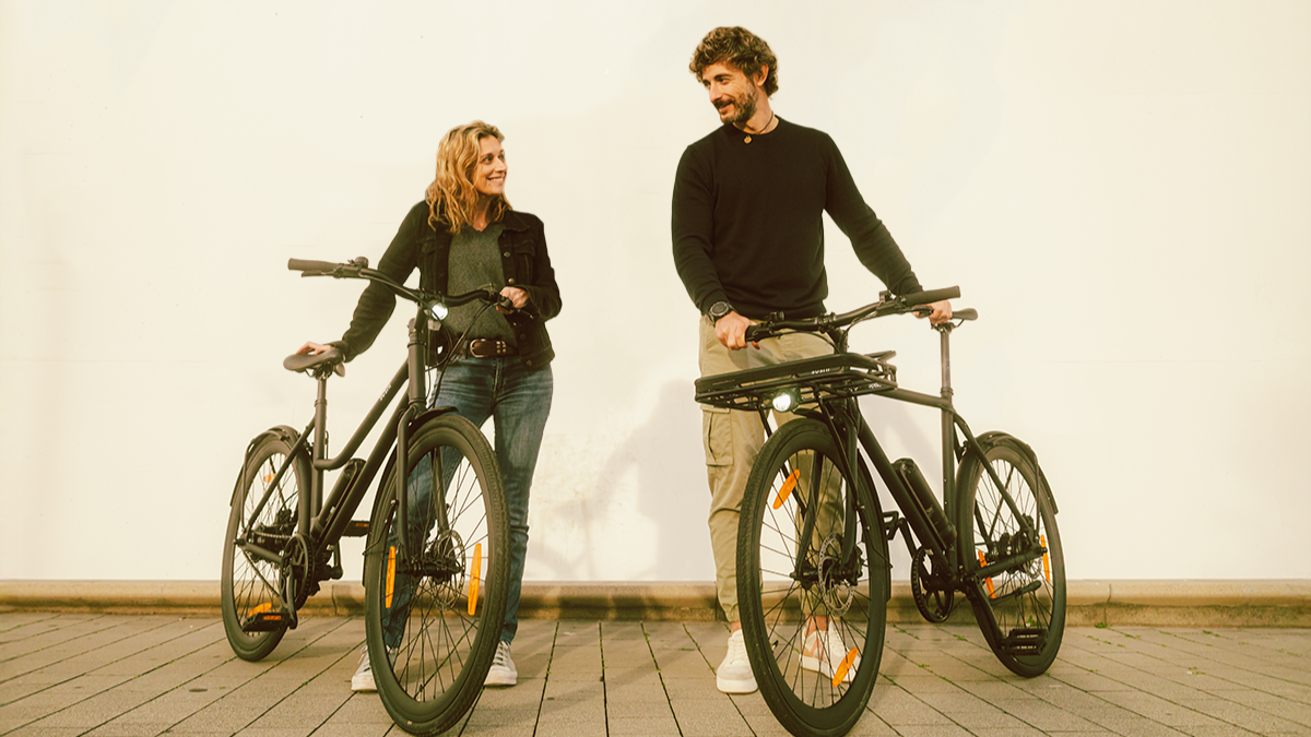 Frau und Mann stehen mit City E-Bike California Roll 3.0+ und Maki 3.0+ in schwarz vor Hauswand